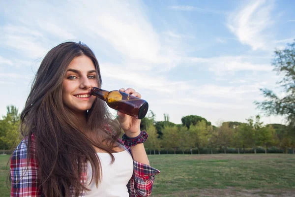 Ganska ung brunett med blå ögon dricka ur en flaska öl i parken. — Stockfoto