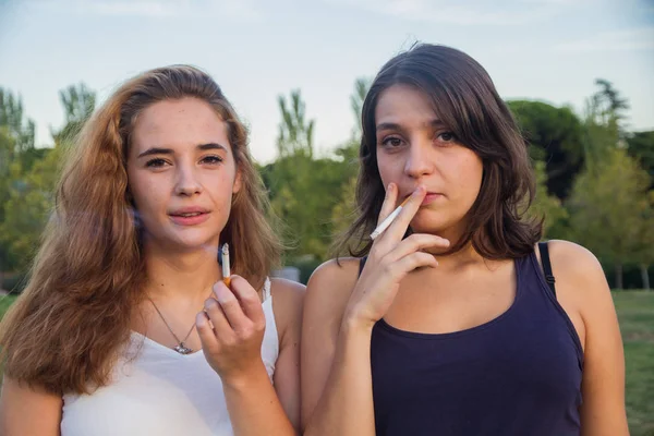 Dos amigas fumando cigarrillos en el parque en un día nublado. Son de celebración y son felices . — Foto de Stock