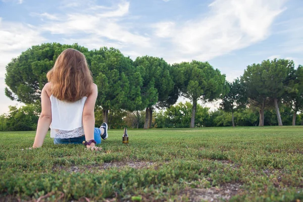 Menina tem sua garrafa de cerveja no chão, enquanto olha para o horizonte pensativo. Sua mão e cerveja descansando na grama . — Fotografia de Stock