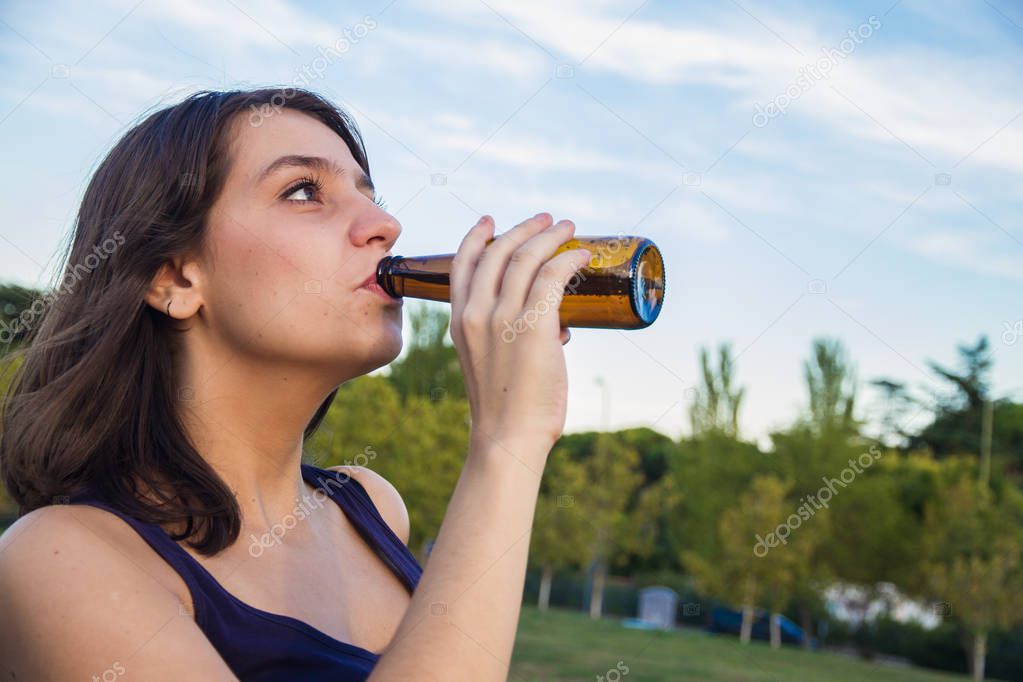 Pretty young brunette drinking from a bottle of beer in the park.