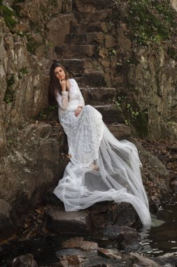 Gelin taş merdivenle kırsalında oturan mavi gözlü genç kadın giyinmiş.