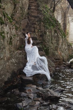 Gelin taş merdivenle kırsalında oturan mavi gözlü genç kadın giyinmiş.