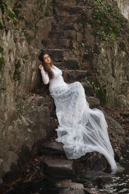 Gelin taş merdivenle kırsalında oturan mavi gözlü genç kadın giyinmiş.