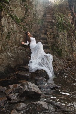 Gelin taş merdivenle kırsalında oturan mavi gözlü genç kadın giyinmiş.