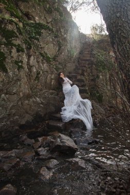 Gelin taş merdivenle kırsalında oturan mavi gözlü genç kadın giyinmiş.