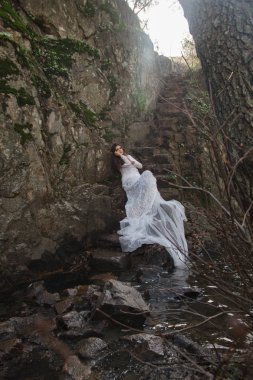 Gelin taş merdivenle kırsalında oturan mavi gözlü genç kadın giyinmiş.