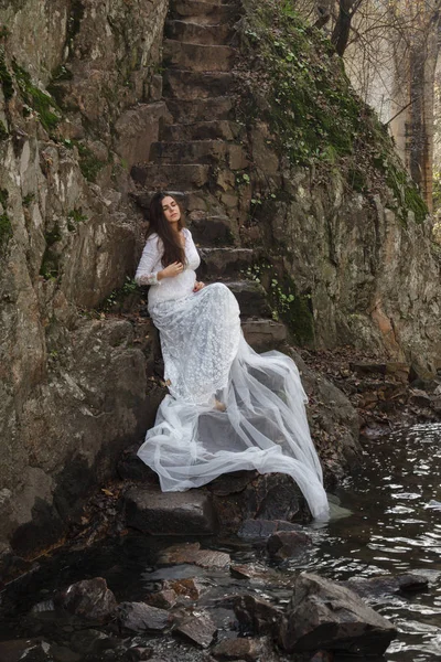Young woman with blue eyes dressed bride sitting on a stone stairs in the countryside. — Stock Photo, Image