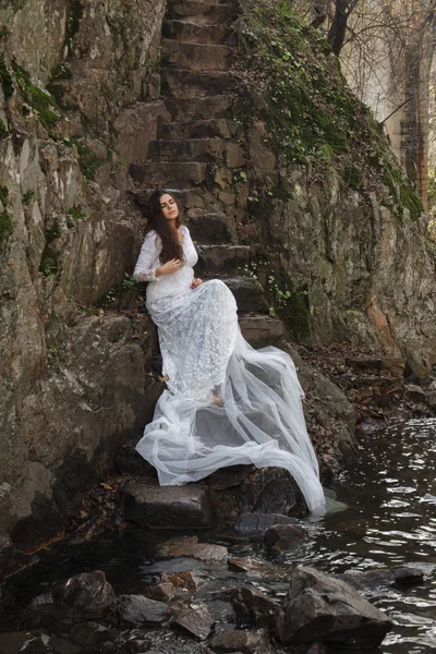 Jeune femme aux yeux bleus mariée habillée assise sur un escalier en pierre dans la campagne . — Photo