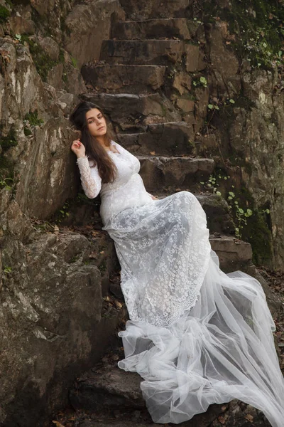 Gelin taş merdivenle kırsalında oturan mavi gözlü genç kadın giyinmiş.