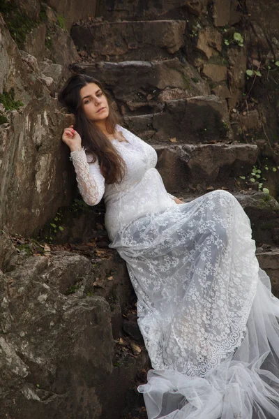 Gelin taş merdivenle kırsalında oturan mavi gözlü genç kadın giyinmiş.