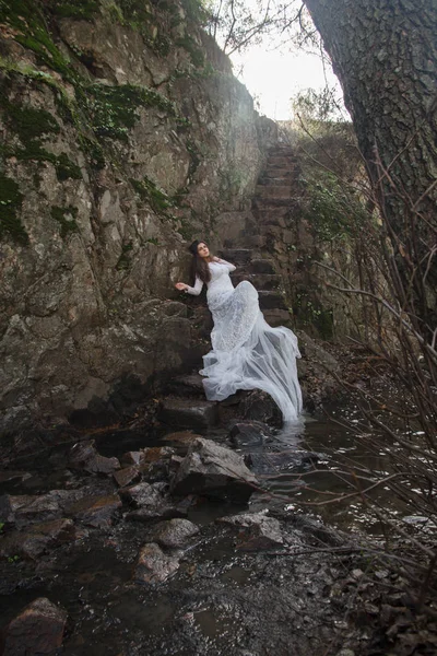 Gelin taş merdivenle kırsalında oturan mavi gözlü genç kadın giyinmiş.