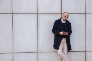 Beyaz duvara yaslanmış ekran dokunarak aşağı seyir bir akıllı telefon tutan genç yakışıklı kel işadamı. 