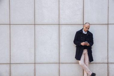 Beyaz duvara yaslanmış ekran dokunarak aşağı seyir bir akıllı telefon tutan genç yakışıklı kel işadamı. 