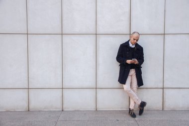 Beyaz duvara yaslanmış ekran dokunarak aşağı seyir bir akıllı telefon tutan genç yakışıklı kel işadamı. 