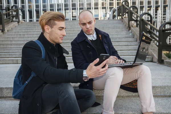 Dos jóvenes emprendedores, uno calvo y el otro rubio trabajando con un portátil y el teléfono inteligente en unas escaleras de la ciudad . —  Fotos de Stock