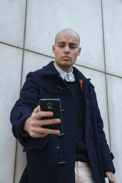 Young handsome bald businessman holding a smart phone, looking down tapping screen leaning against a white wall.