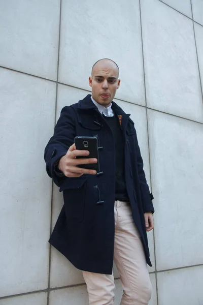 Joven hombre de negocios calvo guapo sosteniendo un teléfono inteligente, mirando hacia abajo golpeando la pantalla apoyada contra una pared blanca . — Foto de Stock