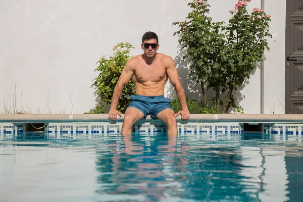 Güneşli bir günde havuz kaldırım kenarında oturan güçlü genç adam 