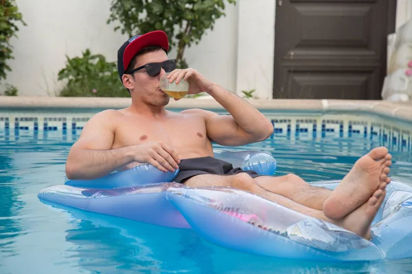 Young strong man drinking beer on an air bed in the pool — Stock Photo, Image