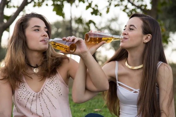 Two young stylish women relaxing together in park and drinking beer looking at each other.
