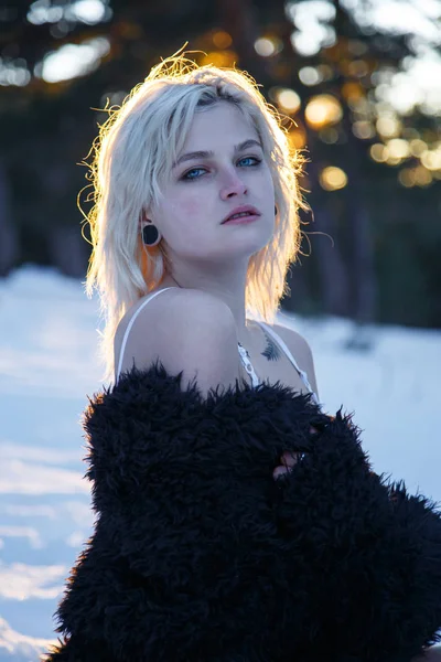 Young blonde woman in dress and fur coat looking at camera in snowy nature.