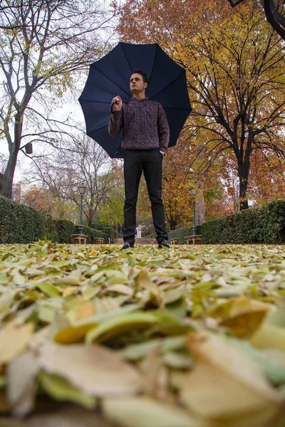 Young man with umbrella walking by fallen leaves on the ground in a park a cloudy day.
