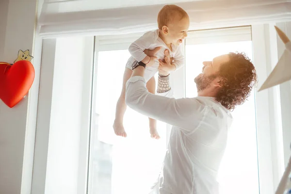Baba, güneşli bir odada pencerenin yanında yeni doğmuş tatlı bir bebekle oynuyor. Sevgi dolu bir baba oğlunu büyütüp gülümsetiyor. Evde güneşli günlerin tadını çıkarıyorum. Mükemmel ebeveynlik ilişkisi