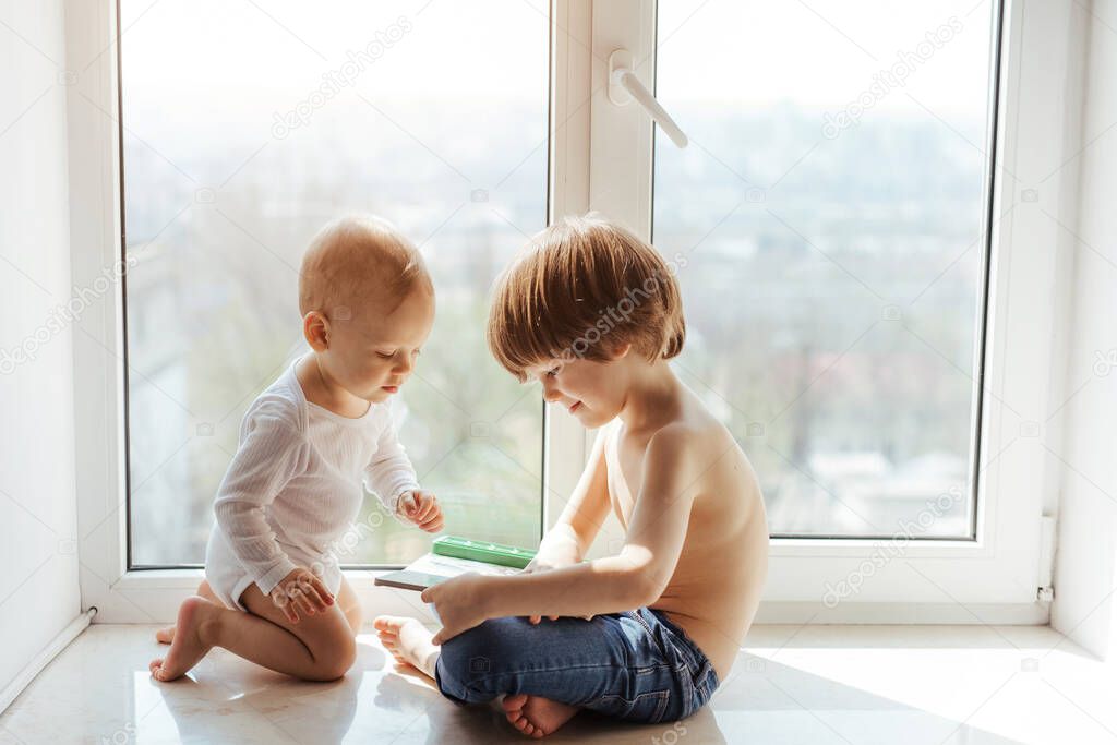 Two little brothers spending time together near window. Older brother reads a book to the little one. True kid friendship. Boys dancing. Kids hugs and kisses