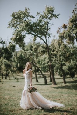 Beyaz modern bir hippi elbisesi içinde yürüyen bir gelin portresi. Günbatımında ormanda eski moda elbiseli beyaz sarışın gelin. Bir buket çiçekle poz veren çekici bir bayan.