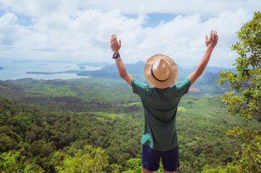 Hayranlıkla ellerini kaldırmış genç bir gezgin yüksek dağın tepesinde duruyor ve orman, tepeler ve denizin manzarasını izliyor. Başarı kavramı, sağlıklı yaşam tarzı, doğayla uyum..