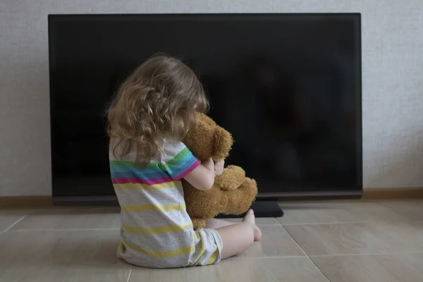 Retrato conceitual. Menina está sentada no chão abraçando com um brinquedo de pelúcia no fundo de uma TV tela preta — Fotografia de Stock