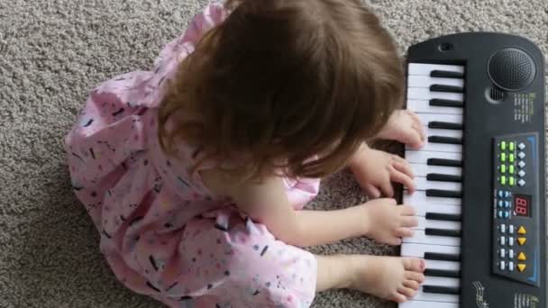 Una niña está sentada en la alfombra y tocando un juguete de piano eléctrico. vista desde arriba. Vídeo Full HD — Vídeos de Stock