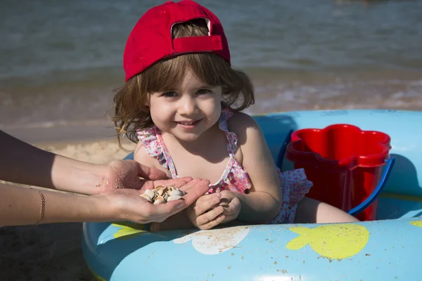 Pequeña linda chica en una gorra roja se sienta en una piscina para niños y juega con su madre en conchas marinas — Foto de Stock