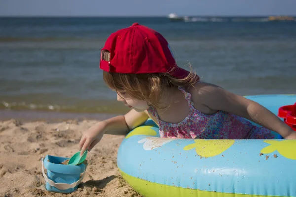 Pequeña linda chica en una gorra roja se sienta en una piscina para niños en la playa — Foto de Stock