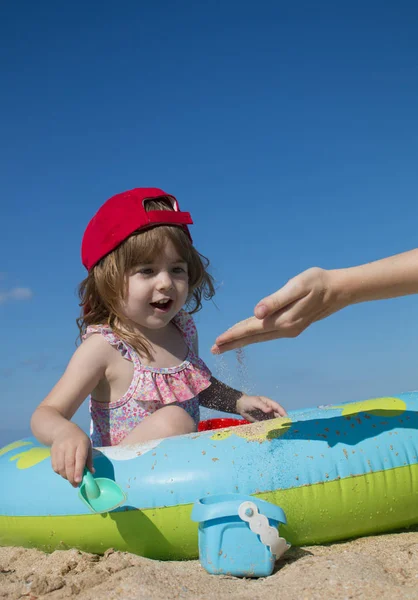 Pequeña linda niña en una gorra roja se sienta en una piscina para niños y juega con su madre con arena — Foto de Stock