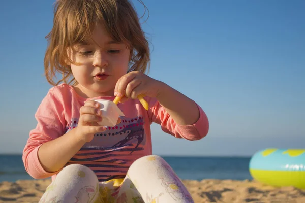 Chica muy bonita comiendo patatas fritas y salsa. chica sentada en la arena en la playa contra el mar . — Foto de Stock