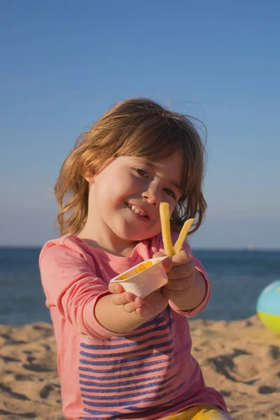 Chica muy linda sostiene en las manos fritas y salsa. chica sentada en la arena en la playa contra el cielo . — Foto de Stock