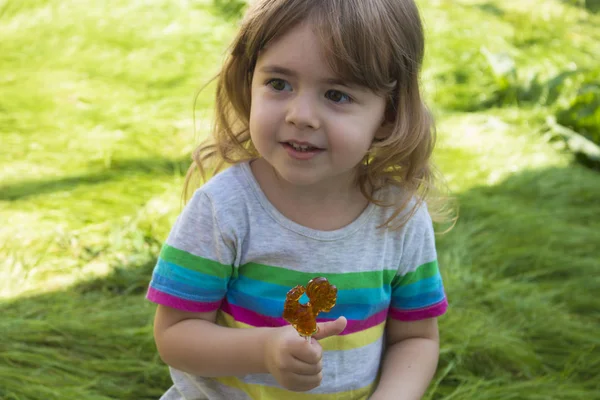 Little pretty girl eating caramel lollipop on a background of green grass and smiling — Stock Photo, Image