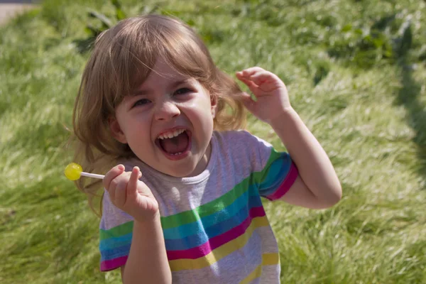 Lilla vackra flicka äta karamell lollipop på en bakgrund av grönt gräs och leende — Stockfoto