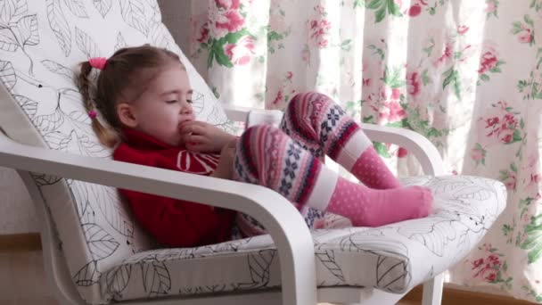 Una linda niña está sentada en un sillón cerca de la ventana jugando con una tableta digital — Vídeo de stock