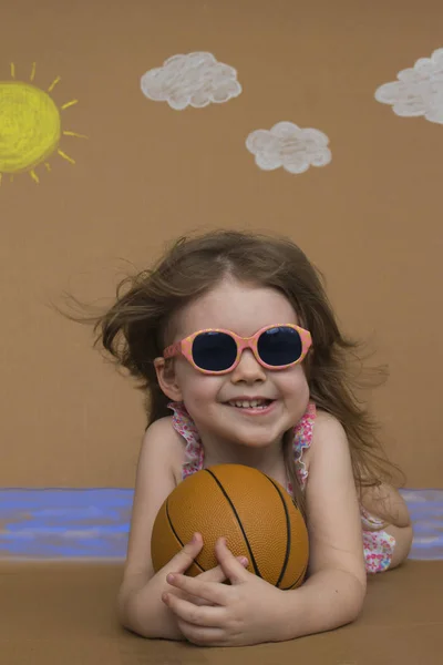 Konceptuella sommarlovet. Söt liten flicka i baddräkt och solglasögon spela med bollen. Bakgrund av målade sol, vita moln och hav. — Stockfoto