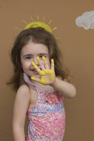 Conceptual summer vacation. Cute little girl in swimsuit with paint on her palm play on background of painted sun and white clouds. — Stock Photo, Image