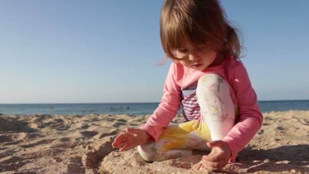 美しい少女は、海と青い空の背景のビーチで砂で遊んでください。 — ストック動画