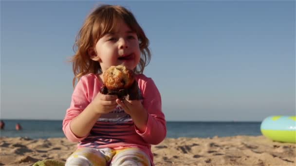 Un niño pequeño y lindo comiendo una magdalena y bebe agua de una botella. Se sienta en la playa contra el mar — Vídeos de Stock