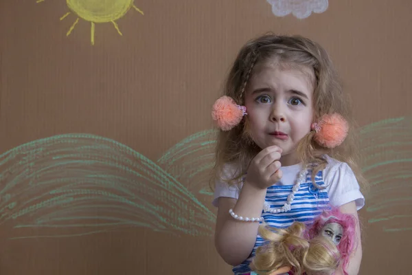 Retrato emocional niña y coletas miran a la cámara. Bebé jugando con su muñeca. fondo de sol pintado y nubes blancas — Foto de Stock