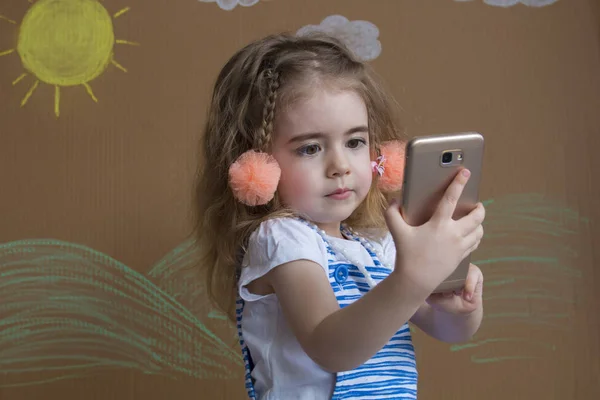 Duygusal portre küçük sarışın kız ve pigtails, bir akıllı tutan el telefon. Arka plan beyaz bulutlar ve boyalı güneş