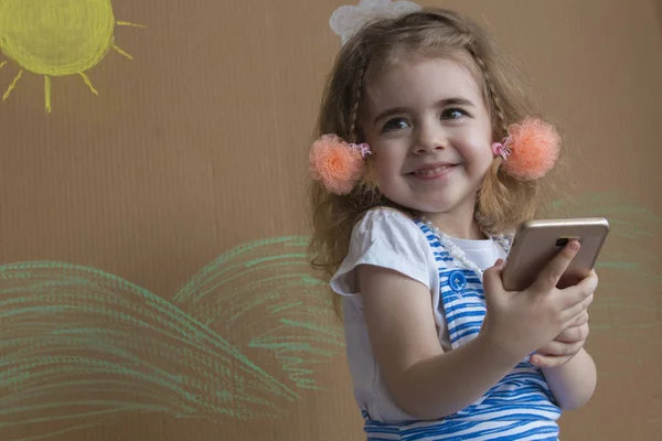 Retrato emocional niña rubia, sosteniendo un teléfono inteligente en la mano y sonriendo. Fondo de sol pintado y nubes blancas — Foto de Stock