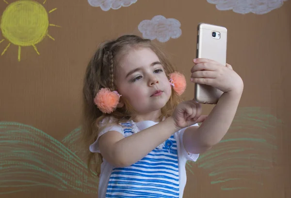 Close-up of a beautiful little girl smiling with a funny face makes selfie on a background in studio — Stock Photo, Image
