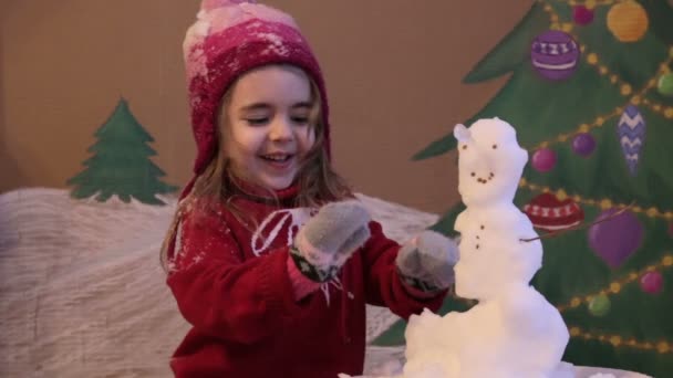 여자는 겨울에는 눈사람을 sculpts. 겨울에 귀여운 여자의 감정적 초상화입니다. 그린 크리스마스 트리와 눈의 배경 — 비디오