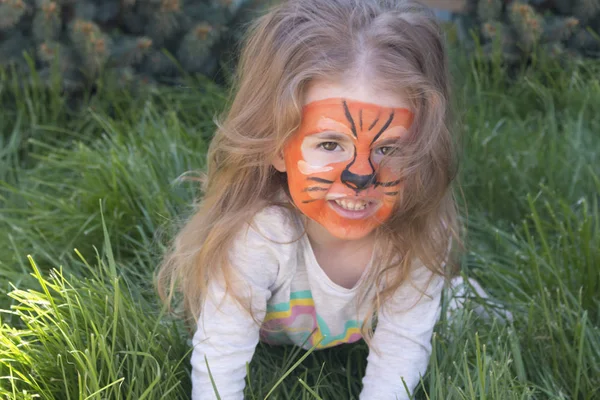 Porträtt av en liten flicka med tiger aqua makeup, leende och sitter i gröna gräset. — Stockfoto